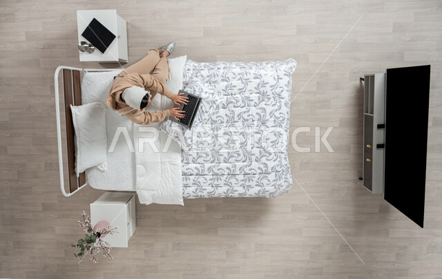 استخدام التقنيه الحديثه والاجهزه الالكترونيه للتواصل عن بعد، الدفع الاكتروني والتسوق باستخدام البطاقات البنكيه، التواصل مع الاهل والاصدقاء باستخدام الجهاز المحمول، بورتريه عامودي علوي لسيدة عربية خليجية سعودية محجبه  تقوم باستخدام الاجهزه الحديثه جالسه على سريرها في غرفه النوم، خلفيه بيج