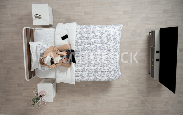 Using modern technology and electronic devices to communicate remotely, electronic payment and shopping using bank cards, communicating with family and friends using a mobile device, a vertical portrait of a veiled Saudi Gulf Arab woman using modern devices, sitting on her bed in the bedroom, beige background.