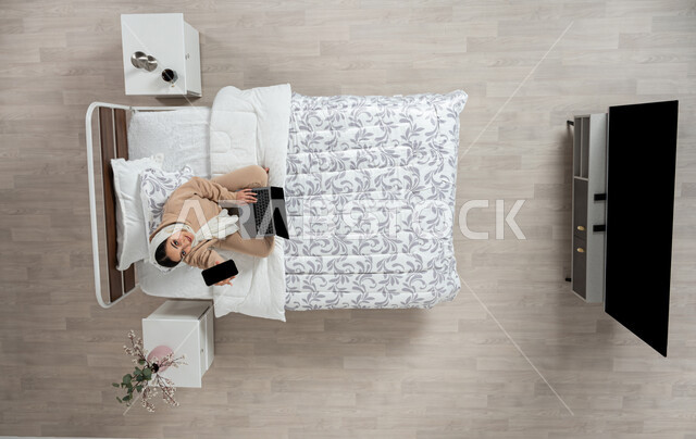 استخدام التقنيه الحديثه والاجهزه الالكترونيه للتواصل عن بعد، الدفع الاكتروني والتسوق باستخدام البطاقات البنكيه، التواصل مع الاهل والاصدقاء باستخدام الجهاز المحمول، بورتريه عامودي علوي لسيدة عربية خليجية سعودية محجبه  تقوم باستخدام الاجهزه الحديثه جالسه على سريرها في غرفه النوم، خلفيه بيج