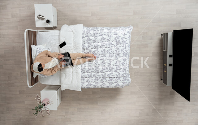 استخدام التقنيه الحديثه والاجهزه الالكترونيه للتواصل عن بعد، الدفع الاكتروني والتسوق باستخدام البطاقات البنكيه، التواصل مع الاهل والاصدقاء باستخدام الجهاز المحمول، بورتريه عامودي علوي لسيدة عربية خليجية سعودية محجبه  تقوم باستخدام الاجهزه الحديثه جالسه على سريرها في غرفه النوم، خلفيه بيج