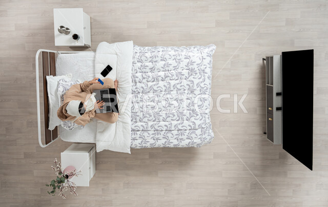 Using modern technology and electronic devices to communicate remotely, electronic payment and shopping using bank cards, communicating with family and friends using a mobile device, a vertical portrait of a veiled Saudi Gulf Arab woman using modern devices, sitting on her bed in the bedroom, beige background.