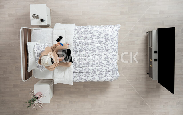 Using modern technology and electronic devices to communicate remotely, electronic payment and shopping using bank cards, communicating with family and friends using a mobile device, a vertical portrait of a veiled Saudi Gulf Arab woman using modern devices, sitting on her bed in the bedroom, beige background.