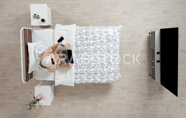 استخدام التقنيه الحديثه والاجهزه الالكترونيه للتواصل عن بعد، الدفع الاكتروني والتسوق باستخدام البطاقات البنكيه، التواصل مع الاهل والاصدقاء باستخدام الجهاز المحمول، بورتريه عامودي علوي لسيدة عربية خليجية سعودية محجبه  تقوم باستخدام الاجهزه الحديثه جالسه على سريرها في غرفه النوم، خلفيه بيج