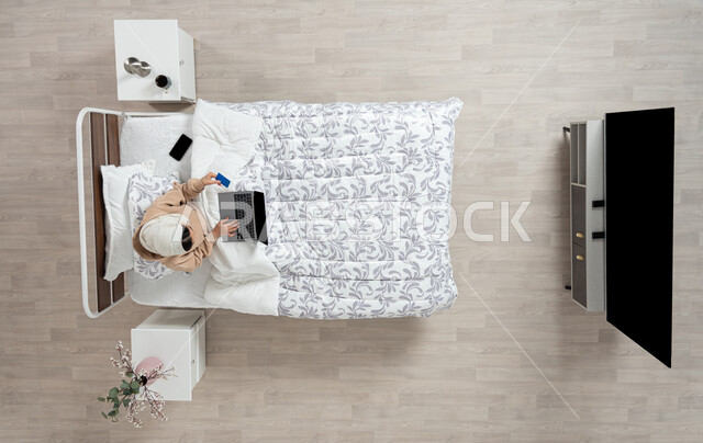 استخدام التقنيه الحديثه والاجهزه الالكترونيه للتواصل عن بعد، الدفع الاكتروني والتسوق باستخدام البطاقات البنكيه، التواصل مع الاهل والاصدقاء باستخدام الجهاز المحمول، بورتريه عامودي علوي لسيدة عربية خليجية سعودية محجبه  تقوم باستخدام الاجهزه الحديثه جالسه على سريرها في غرفه النوم، خلفيه بيج