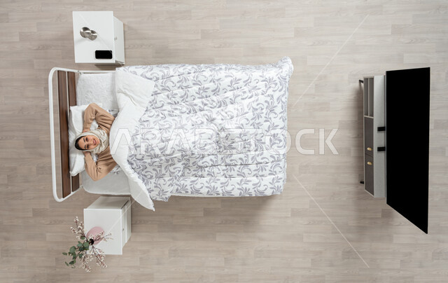 Sleeping and relaxing on a soft bed, feeling happy and relaxed, an overhead portrait of a veiled Saudi Gulf Arab woman lying in her bed under the quilt, placing her hands behind her head, looking up with a smile, gesture movements, different facial and hand expressions, beige background