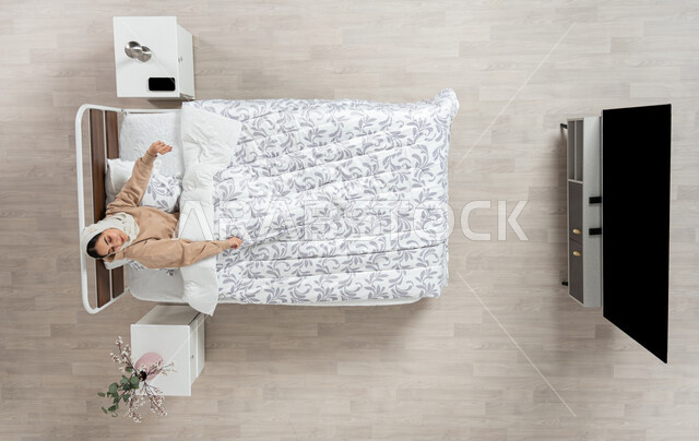 رفع اليدين عند الاستيقاظ، بورتريه علوي عامودي لسيده عربيه خليجيه سعوديه محجبه  مستلقيه في سريرها تقوم بالتمطع، التمطيع والتمطيط، مفهوم تمدد الجسم والاستعداد للنشاط والحركه، بدايه يوم جديد، خلفيه بيج