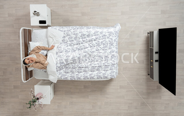 رفع اليدين للأعلى بإيماءات التعب والارهاق، بورتريه عامودي علوي لامرأة عربيه خليجيه سعوديه محجبه ترتدي العباءة الملونة مستلقيه على سرير مريح تقوم بالتثاؤب، تعابير الخمول و الشعور بالنعاس والحاجه الى الراحة و النوم، خلفية بيج