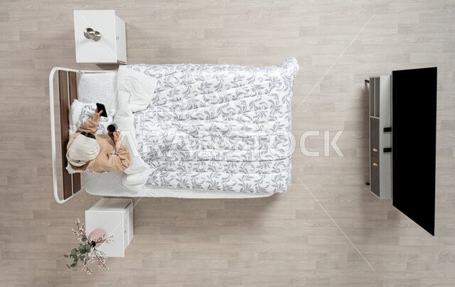 ايماءات تدل على السعادة والاستمتاع بإجراء مكالمة فيديو, بورتريه عامودي علوي لإمرأة عربية خليجية سعودية محجبه تجلس في سريرها مستلقيه تحت لحافها تحمل بيدها كوب القهوة وتستخدم الهاتف المحمول ، استخدام البرامج والتطبيقات الالكترونية، الاستمتاع بالمشروبات الساخنه والمنبهات، خلفيه بيج