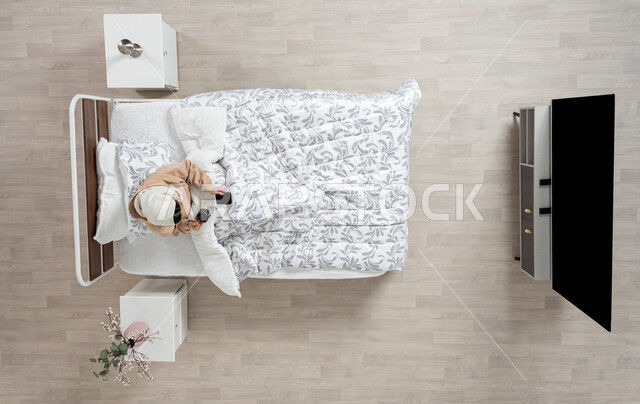 ايماءات تدل على السعادة والاستمتاع بإجراء مكالمة فيديو, بورتريه عامودي علوي لإمرأة عربية خليجية سعودية محجبه تجلس في سريرها مستلقيه تحت لحافها تحمل بيدها كوب القهوة وتستخدم الهاتف المحمول ، استخدام البرامج والتطبيقات الالكترونية، الاستمتاع بالمشروبات الساخنه والمنبهات، خلفيه بيج