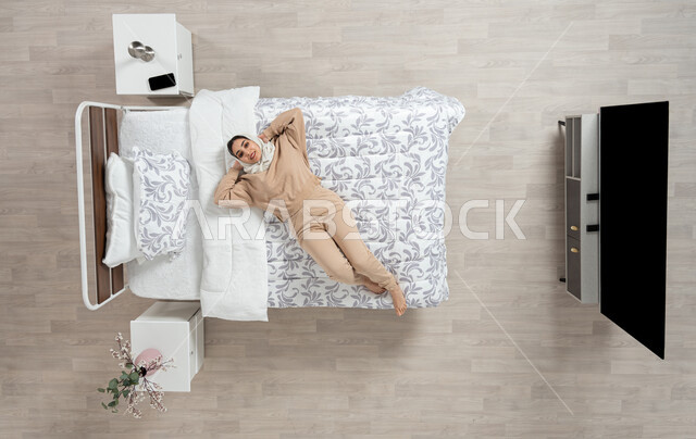 النوم والاسترخاءعلى سرير ناعم، الاحساس بالسعاده والاستجمام، بورتريه عامودي علوي لسيده عربيه خليجيه سعوديه محجبه ترتدي الزي الكاجوال متمدده على سريرها بشكل عامودي مائل،  حركات وايماءات وتعابير وجه ويدين مختلفه، خلفيه بيج