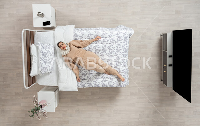 النوم والاسترخاءعلى سرير ناعم، الاحساس بالسعاده والاستجمام، بورتريه عامودي علوي لسيده عربيه خليجيه سعوديه محجبه ترتدي الزي الكاجوال متمدده على سريرها بشكل عامودي مائل،  حركات وايماءات وتعابير وجه ويدين مختلفه، خلفيه بيج