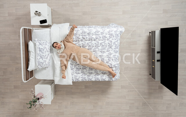 النوم والاسترخاءعلى سرير ناعم، الاحساس بالسعاده والاستجمام، بورتريه عامودي علوي لسيده عربيه خليجيه سعوديه محجبه ترتدي الزي الكاجوال متمدده على سريرها بشكل عامودي مائل، حركات وايماءات وتعابير وجه ويدين مختلفه، خلفيه بيج