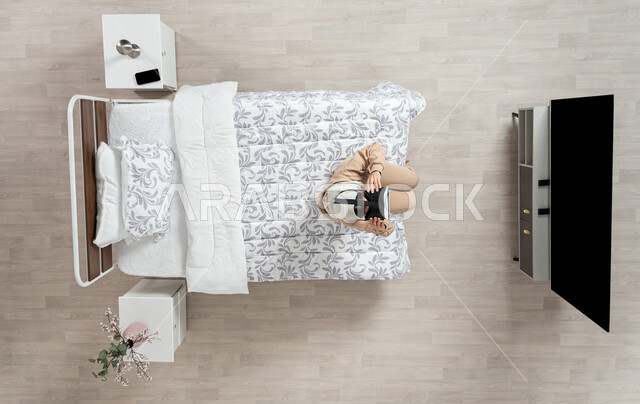 الاندماج والتركيز في الواقع الافتراضي، محاكاة العالم الخيالي، بورتريه عامودي علوي لسيده عربية خليجية سعودية محجبه تجلس على سريرها تنظر الى التلفاز من خلال نظارات الواقع الافتراضي (الفي ار) ممسكه بها بيديها، التحول الرقمي والتقدم التقني، خلفيه بيج