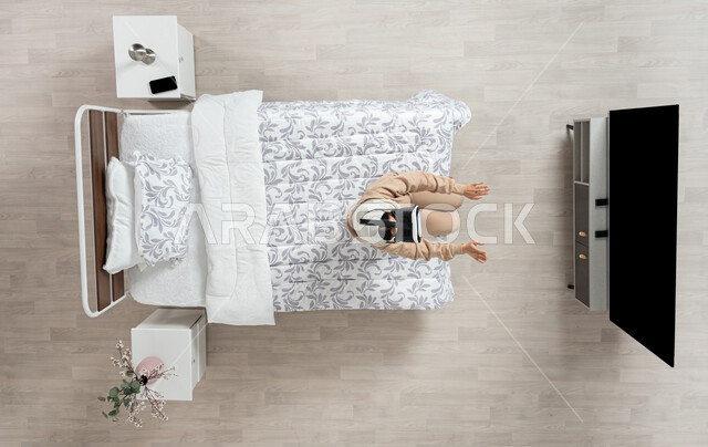 ايماءات الاندماج والتركيز في الواقع الافتراضي، محاكاة العالم الخيالي، بورتريه عامودي علوي لسيده عربية خليجية سعودية محجبه تجلس على سريرها تمد يديها الى الامام، تنظر الى التلفاز من خلال نظارات الواقع الافتراضي (الفي ار)، التحول الرقمي والتقدم التقني، خلفيه بيج