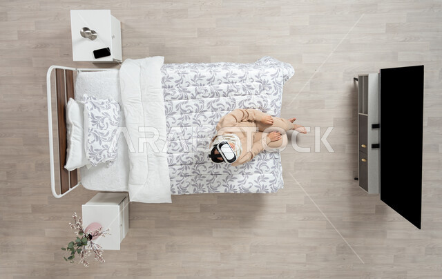 الاندماج والتركيز في الواقع الافتراضي، محاكاة العالم الخيالي،  بورتريه عامودي علوي لسيده عربية خليجية سعودية محجبه تجلس على سريرها تضع يديها على ركبتيها وتنظر الى التلفاز من خلال نظارات الواقع الافتراضي (الفي ار)، التحول الرقمي والتقدم التقني، خلفيه بيج