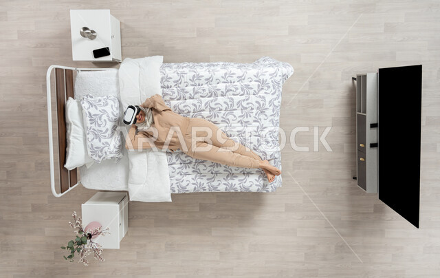 محاكاه العالم الخيالي، بورتريه عامودي علوي لسيده عربيه خليجيه سعوديه ترتدي نظارات (الفي ار) مستلقيه على ظهرها، أمراه مبتسمه تنظر من خلال نظارات الواقع الافتراضي الى الاعلى تضع يديها خلف رأسها وتقوم بضم ساقيها على سريرها، خلفيه بيج