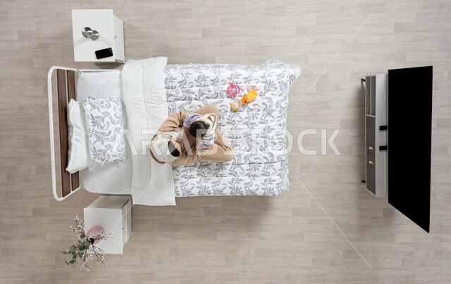 النظر إلى الاطفال بإيماءات الحب والحنان، مداعبه الام لاطفالها الصغار، بورتريه عامودي علوي لأم عربيه خليجيه سعوديه  محجبة تحمل وتداعب ابنتها حديثه الولاده على سريرها, طفله ترتدي لباسا قطنيا ناعم، الرعاية والاهتمام بالأبناء, خلفيه بيج