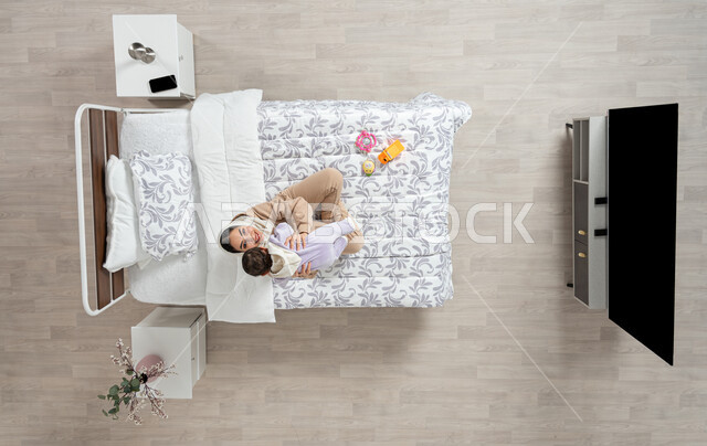 النظر إلى الاطفال بإيماءات الحب والحنان، مداعبه الام لاطفالها الصغار، النظر الى الاعلى باتسامه وسعاده، بورتريه عامودي علوي لأم عربيه خليجيه سعوديه  محجبة تحمل وتداعب ابنتها حديثه الولاده على سريرها, الرعاية والاهتمام بالأبناء, خلفيه بيج