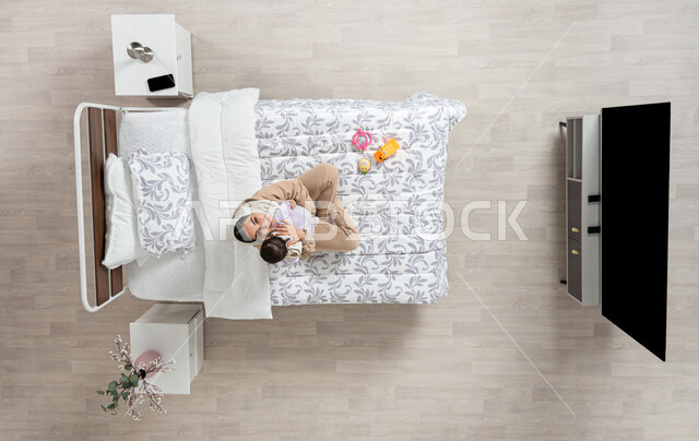 Looking at children with gestures of love and tenderness, mother caressing her young children, looking up with a smile and happiness, vertical portrait of a veiled Saudi Arabian Gulf mother holding and caressing her newborn daughter on her bed, care and attention for children, beige background