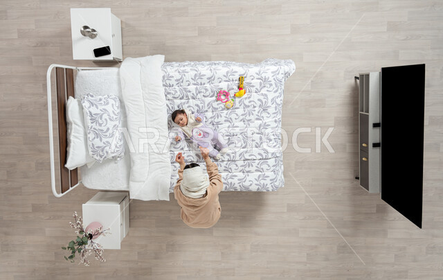 النظر إلى الاطفال بإيماءات الحب والحنان، مداعبه الام لاطفالها الصغار، بورتريه عامودي علوي لأم عربيه خليجيه سعوديه  محجبة تلعب مع ابنتها حديثه الولاده على سريرها, رضيعه ترتدي بربتوز قطني ناعم, الرعاية والاهتمام بالأبناء, خلفيه بيج