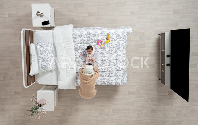 النظر إلى الاطفال بإيماءات الحب والحنان، مداعبه الام لاطفالها الصغار، بورتريه عامودي علوي لأم عربيه خليجيه سعوديه  محجبة تلعب مع ابنتها حديثه الولاده على سريرها, رضيعه ترتدي بربتوز قطني ناعم, الرعاية والاهتمام بالأبناء, خلفيه بيج