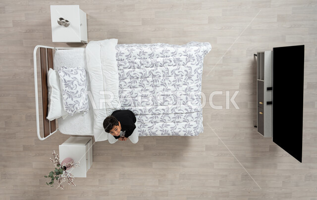 الاسترخاء بالجلوس والتأمل، بورتريه علوي عامودي لولد عربي خليجي سعودي يرتدي الزي الكاجوال يجلس على حافه السرير في غرفه نومه، ينظر إلى الأعلى بإيماءات الفرح والسعادة، خلفية بيج