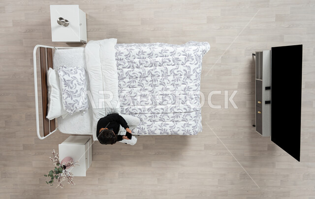 استعمال الأجهزة الإلكترونية الحديثة، التواصل مع الاهل والاصدقاء ، بورتريه عامودي علوي لولد عربي خليجي سعودي يجلس على سريره يمسك بيده جهازه المحمول، تلميذ يرتدي الزي الكاجوال، اجراء المحادثات والشات عن طريق الاونلاين، خلفيه بيج