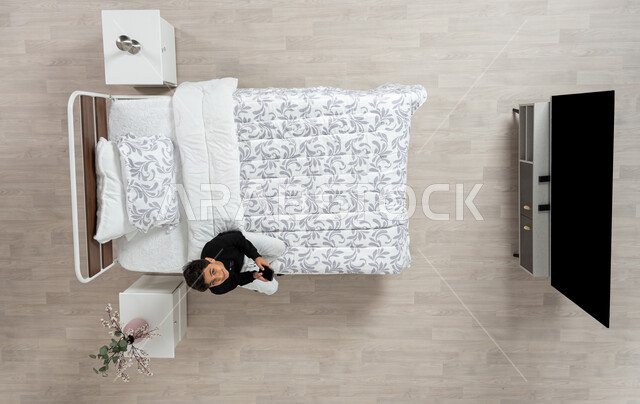 استعمال الأجهزة الإلكترونية الحديثة، التواصل مع الاهل والاصدقاء ، بورتريه عامودي علوي لولد عربي خليجي سعودي مبتسم  يجلس على سريره ينظر الى الاعلى و يمسك بيده جهازه المحمول، تلميذ يرتدي الزي الكاجوال، خلفيه بيج