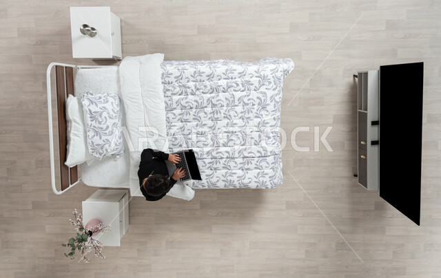 استعمال الأجهزة الإلكترونية الحديثة، التواصل مع الاهل والاصدقاء ، بورتريه عامودي علوي لولد عربي خليجي سعودي يجلس على سريره يمسك بيده جهاز الاب توب المحمول، تلميذ يرتدي الزي الكاجوال، تصفح المواقع الاكترونيه والاونلاين، الدردشه والشات الاكتروني، خلفيه بيج