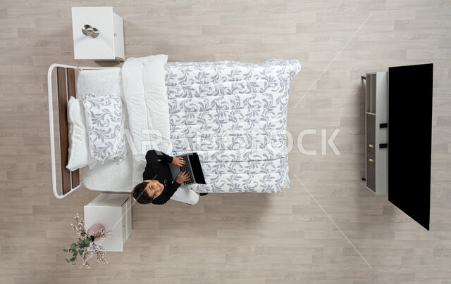 استعمال الأجهزة الإلكترونية الحديثة، التواصل مع الاهل والاصدقاء ، بورتريه عامودي علوي لولد عربي خليجي سعودي يجلس على سريره يمسك بيده جهاز الاب توب المحمول، تلميذ يرتدي الزي الكاجوال ينظر الى الاعلى بابتسامه، تصفح المواقع الاكترونيه والاونلاين، الدردشه والشات الاكتروني، خلفيه بيج