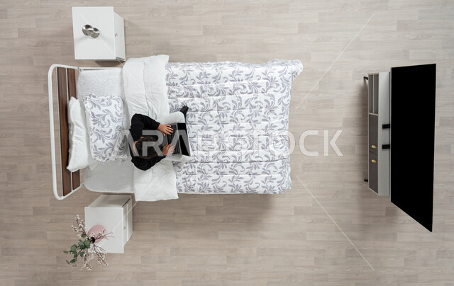 استعمال الأجهزة الإلكترونية الحديثة، التواصل مع الاهل والاصدقاء ، بورتريه عامودي علوي لولد عربي خليجي سعودي يجلس على سريره يمسك بيده جهاز الاب توب المحمول، تلميذ يرتدي الزي الكاجوال، تصفح المواقع الاكترونيه والاونلاين، الدردشه والشات الاكتروني، خلفيه بيج