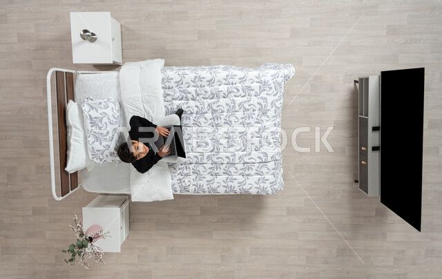 استعمال الأجهزة الإلكترونية الحديثة، التواصل مع الاهل والاصدقاء ، بورتريه عامودي علوي لولد عربي خليجي سعودي يجلس على سريره يمسك بيده جهاز الاب توب المحمول، تلميذ يرتدي الزي الكاجوال ينظر الى الاعلى بابتسامه، تصفح المواقع الاكترونيه والاونلاين، الدردشه والشات الاكتروني، خلفيه بيج