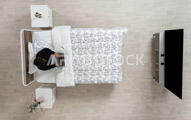 استعمال الأجهزة الإلكترونية الحديثة، التواصل مع الاهل والاصدقاء ، بورتريه عامودي علوي لولد عربي خليجي سعودي يجلس على سريره يمسك بيده جهاز الاب توب المحمول، تلميذ يرتدي الزي الكاجوال، تصفح المواقع الاكترونيه والاونلاين، الدردشه والشات الاكتروني، خلفيه بيج