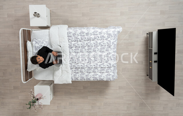 استعمال الأجهزة الإلكترونية الحديثة، التواصل مع الاهل والاصدقاء ، بورتريه عامودي علوي لولد عربي خليجي سعودي يجلس على سريره يمسك بيده جهاز الاب توب المحمول، تلميذ يرتدي الزي الكاجوال ينظر الى الاعلى بابتسامه، تصفح المواقع الاكترونيه والاونلاين، الدردشه والشات الاكتروني، خلفيه بيج