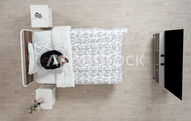 استعمال الأجهزة الإلكترونية الحديثة، التواصل مع الاهل والاصدقاء ، بورتريه عامودي علوي لولد عربي خليجي سعودي يجلس على سريره يمسك بيده جهازه المحمول، تلميذ يرتدي الزي الكاجوال، تصفح المواقع الاكترونيه والاونلاين، الدردشه والشات الاكتروني، خلفيه بيج