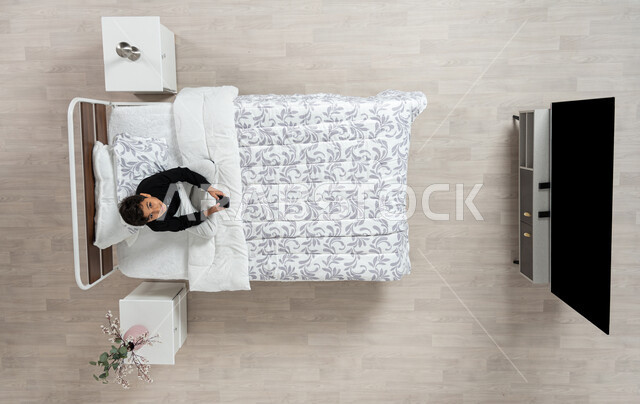 استعمال الأجهزة الإلكترونية الحديثة، التواصل مع الاهل والاصدقاء ، بورتريه عامودي علوي لولد عربي خليجي سعودي مبتسم  يجلس على سريره ينظر الى الاعلى و يمسك بيده جهازه المحمول، تلميذ يرتدي الزي الكاجوال، خلفيه بيج