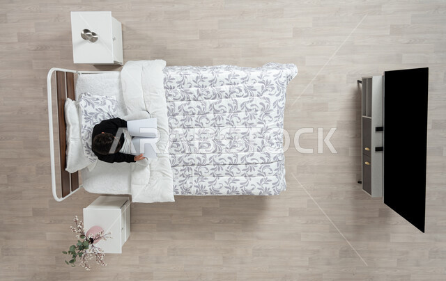 القراءة و الإطلاع و الاستعداد للعالم الدراسي الجديد، صورة علوية عاموديه لولدعربي خليجي سعودي يقوم بقراءة كتاب جالسا على سريره مرتديا الزي الكاجوال، كتب علمية وروايات ثقافية، تنمية المهارات الذهنية و ممارسة هواية القراءه، خلفيه بيج