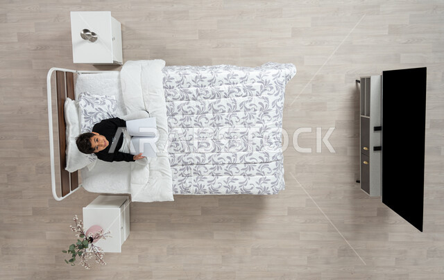 Reading, learning and preparing for the new school world, vertical overhead photo of a Saudi Arabian Gulf boy reading a book sitting on his bed wearing casual clothes looking up smiling, scientific books and cultural novels, developing mental skills and practicing the hobby of reading, beige background