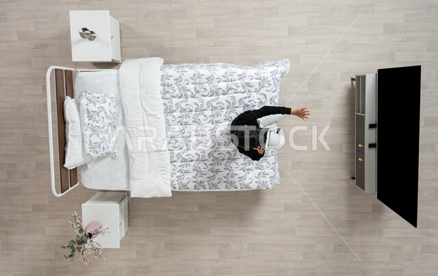 التطور التقني والتكنولوجي، مفهوم التفاعل و الاندماج، بورتريه عامودي علوي  لشاب عربي سعودي خليجي يرتدي الزي الكاجوال ونظارة الواقع الافتراضي ثلاثية الأبعاد يجلس على سريره يقوم بتحريك يده يحاول لمس شيء ما، المتعة بمشاهدة الأفلام و محاكاة العالم الخيالي، خلفيه بيج