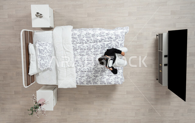 Technical and technological development, the concept of interaction and integration, vertical portrait of a young Saudi Arabian Gulf man wearing casual clothes and 3D virtual reality glasses sitting on his bed moving his hand trying to touch something, enjoy watching movies and simulating the imaginary world, beige background