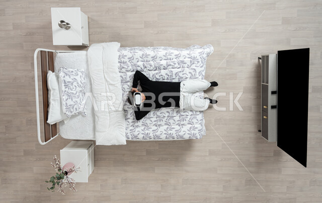 Technical and technological development, the concept of interaction and integration, vertical overhead portrait of a young Saudi Arabian Gulf man wearing casual clothes and 3D virtual reality glasses lying on his bed, enjoyment of watching movies and simulating the imaginary world, beige background