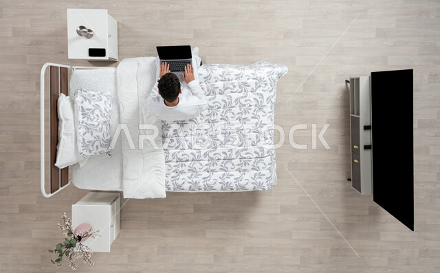 أداء الواجبات والعمل عن بعد، استخدام جهاز الاب التوب في التصفح والدردشه ، بورتريه علوي عامودي لشاب عربي خليجي سعودي يرتدي الثوب التقليدي الابيض يعمل على جهاز الكمبيوتر المحمول جالسا على سريره في غرفه نومه، خلفية بيج