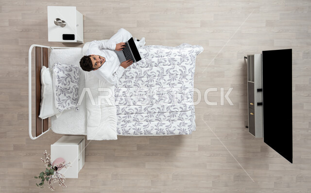 أداء الواجبات والعمل عن بعد ، استخدام جهاز الاب التوب في التصفح والدردشه ، بورتريه علوي عامودي لشاب عربي خليجي سعودي مبتسم ينظر الى الاعلى  يرتدي الثوب التقليدي الابيض يعمل على جهاز الكمبيوتر المحمول جالسا على سريره، خلفية بيج