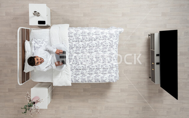 أداء الواجبات والعمل عن بعد ، استخدام جهاز الاب التوب في التصفح والدردشه ، بورتريه علوي عامودي لشاب عربي خليجي سعودي مبتسم ينظر الى الاعلى  يرتدي الثوب التقليدي الابيض يعمل على جهاز الكمبيوتر المحمول جالسا على سريره، خلفية بيج