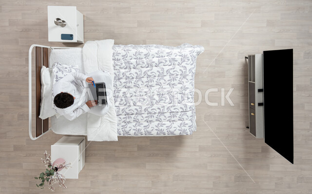 أداء الواجبات والعمل عن بعد، استخدام جهاز الاب التوب في التصفح والدردشه ، بورتريه علوي عامودي لشاب عربي خليجي سعودي يرتدي الثوب التقليدي الابيض يعمل على جهاز الكمبيوتر المحمول جالسا على سريره في غرفه نومه، خلفية بيج