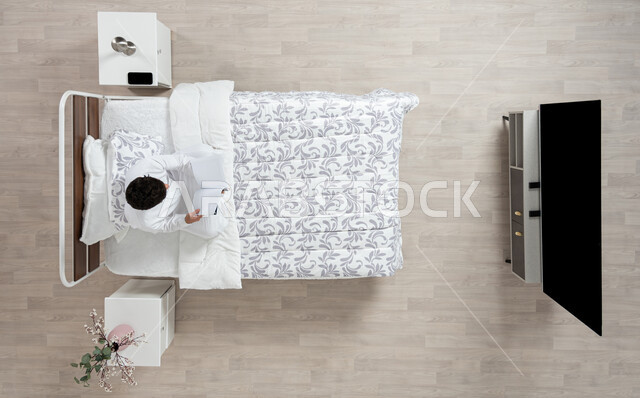 القراءة و الإطلاع و الاستعداد للعالم الدراسي الجديد، صورة علوية عاموديه لشاب عربي خليجي سعودي يقوم بقراءة كتاب جالسا على سريره مرتديا الثوب الابيض التقليدي، كتب علمية وروايات ثقافية، تنمية المهارات الذهنية و ممارسة هواية القراءه، خلفيه بيج