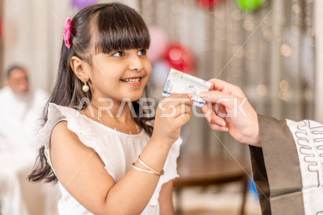 طفلة سعودية خليجية سعيدة ترتدي لباس العيد الانيق ، تأخذ العيدية  ، الاحتفال بعيد الفطر السعيد ، عيد الفطر المبارك  ،  تقديم العيديات و الحلوى ، القرقيعان