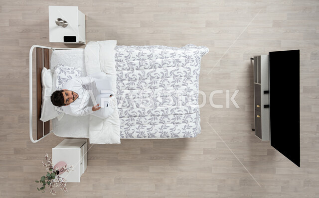 Reading, learning, and preparing for the new academic world, a vertical overhead picture of a Saudi Gulf Arab young man reading a book sitting on his bed, wearing the traditional white dress, looking up with a smile, scientific books and cultural novels, developing mental skills and practicing the hobby of reading, beige background.