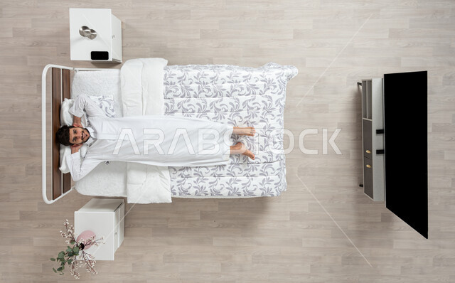 الاسترخاء والراحة وقت القيلوله، بورتريه علوي عامودي لشاب عربي خليجي سعودي يرتدي الثوب الابيض التقليدي مستلقي على سريره واضعا يديه خلف رأسه ينظر الى الكاميرا بابتسامه، ايماءات وجه ويدين تدل على السعادة، الاستمتاع بنوم هادئ وصحي، وسادة بيضاء ناعمة، خلفيه بيج