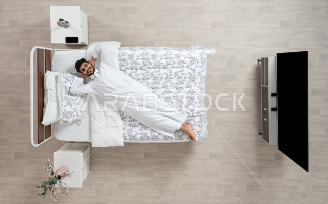 الاسترخاء والراحة وقت القيلوله، بورتريه علوي عامودي لشاب عربي خليجي سعودي يرتدي الثوب الابيض التقليدي مستلقي على سريره واضعا يديه خلف رأسه ينظر الى الكاميرا بابتسامه، ايماءات وجه ويدين تدل على السعادة، الاستمتاع بنوم هادئ وصحي، وسادة بيضاء ناعمة، خلفيه بيج